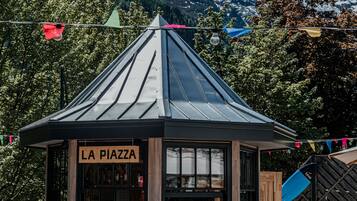 Quiosco de madera llamado "LA PIAZZA" con techo inclinado, rodeado de mesas al aire libre y vistas de montañas nevadas.