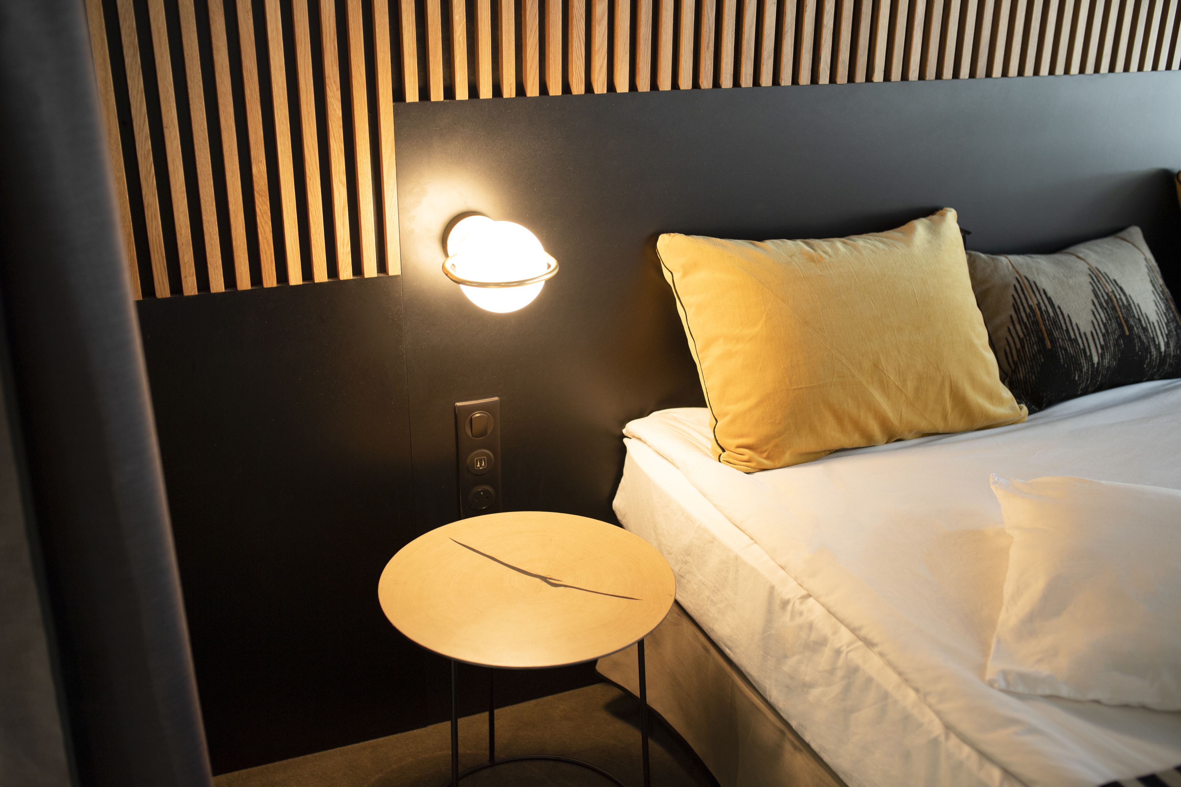 Ein Hotelzimmer mit einem Bett, einem Nachttisch, einer Wandleuchte und einem Holzschrankgitter als Kopfteil.