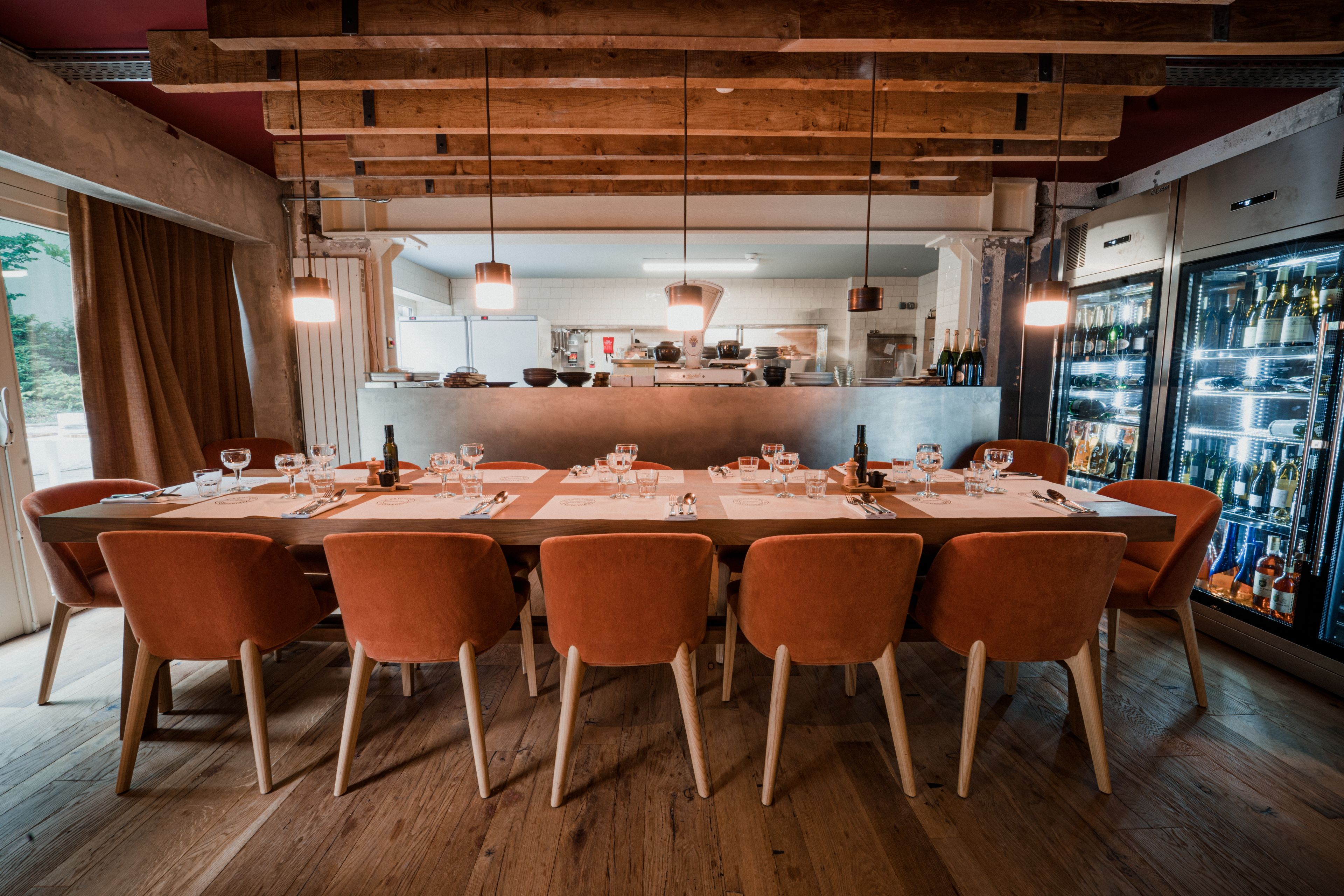 Un restaurant avec une longue table en bois prévue pour six personnes, des chaises orange et un coin bar doté d’un réfrigérateur rempli de bouteilles.