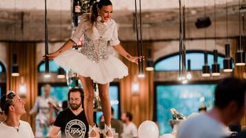 A person in a white dress with a sequined corset standing on a platform, surrounded by people in a room with wooden walls and hanging lights.