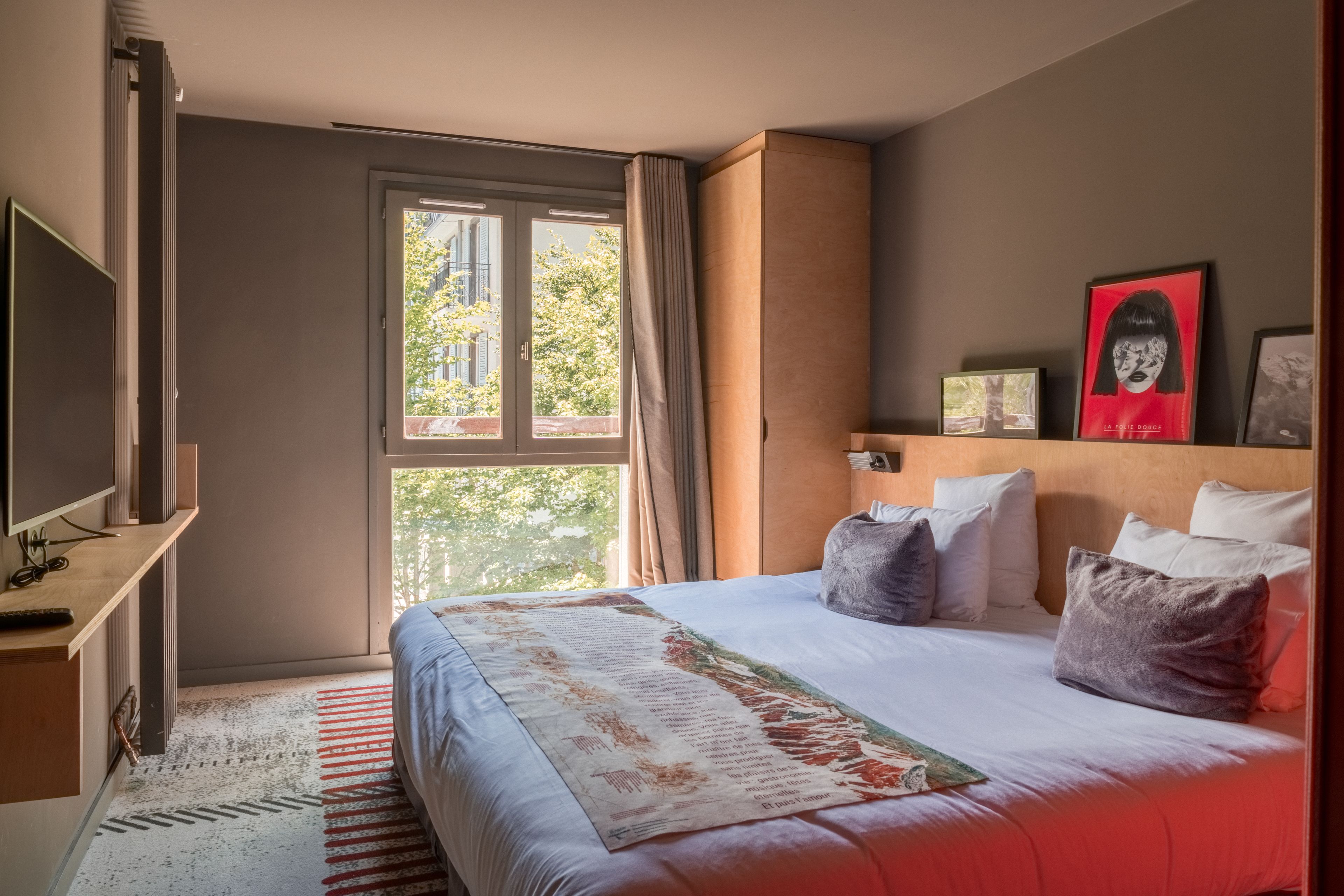 A hotel room with a large bed, a TV, a window with curtains, and a view of trees outside.