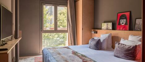 A hotel room with a large bed, a TV, a window with curtains, and a view of trees outside.