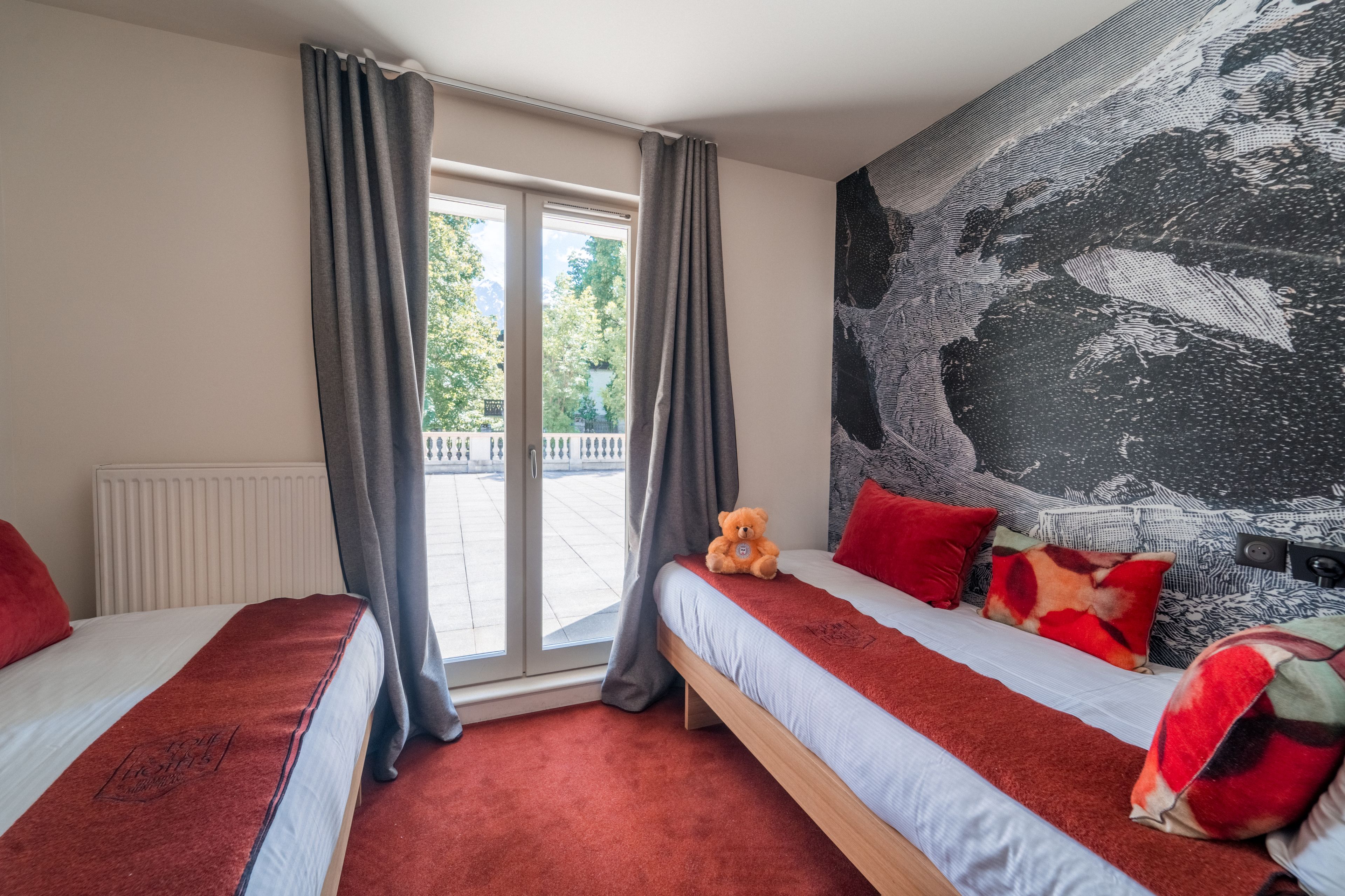 Habitación de hotel con dos camas, un ventanal grande con cortinas, un osito de peluche sobre una cama y un cuadro abstracto de gran tamaño en la pared.