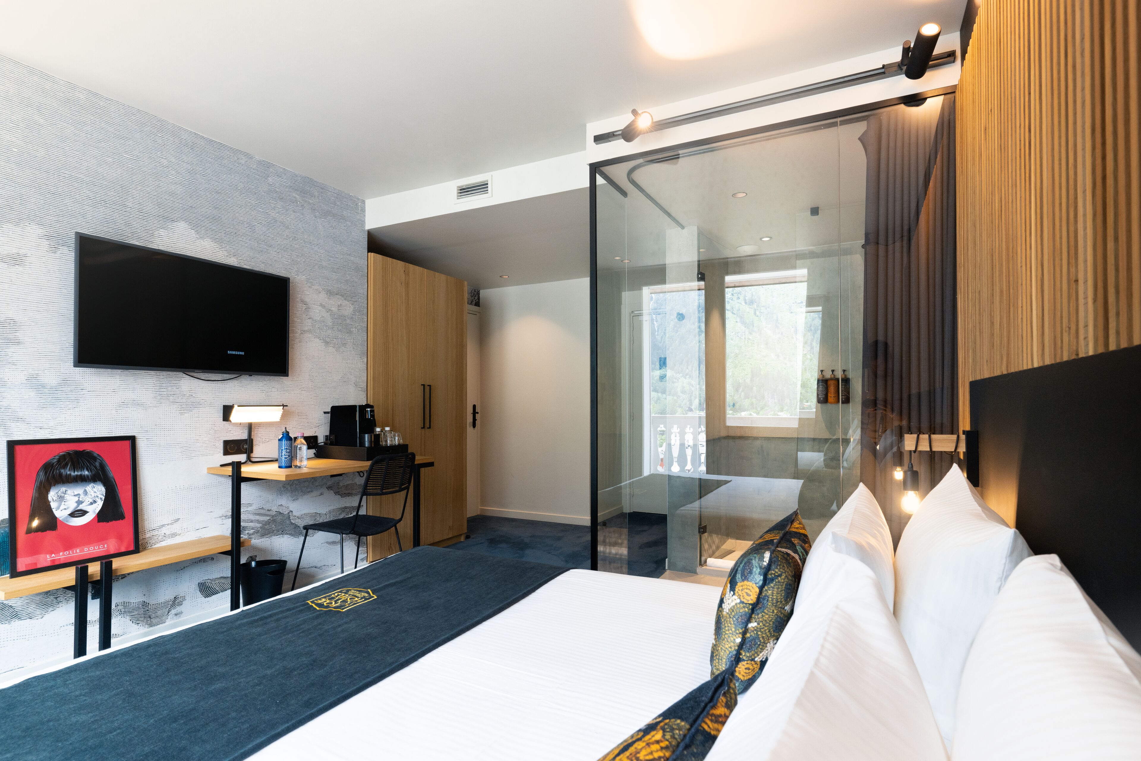 A modern hotel room with a large bed, a desk with a computer, a flat-screen TV mounted on the wall, and a glass partition leading to a balcony.