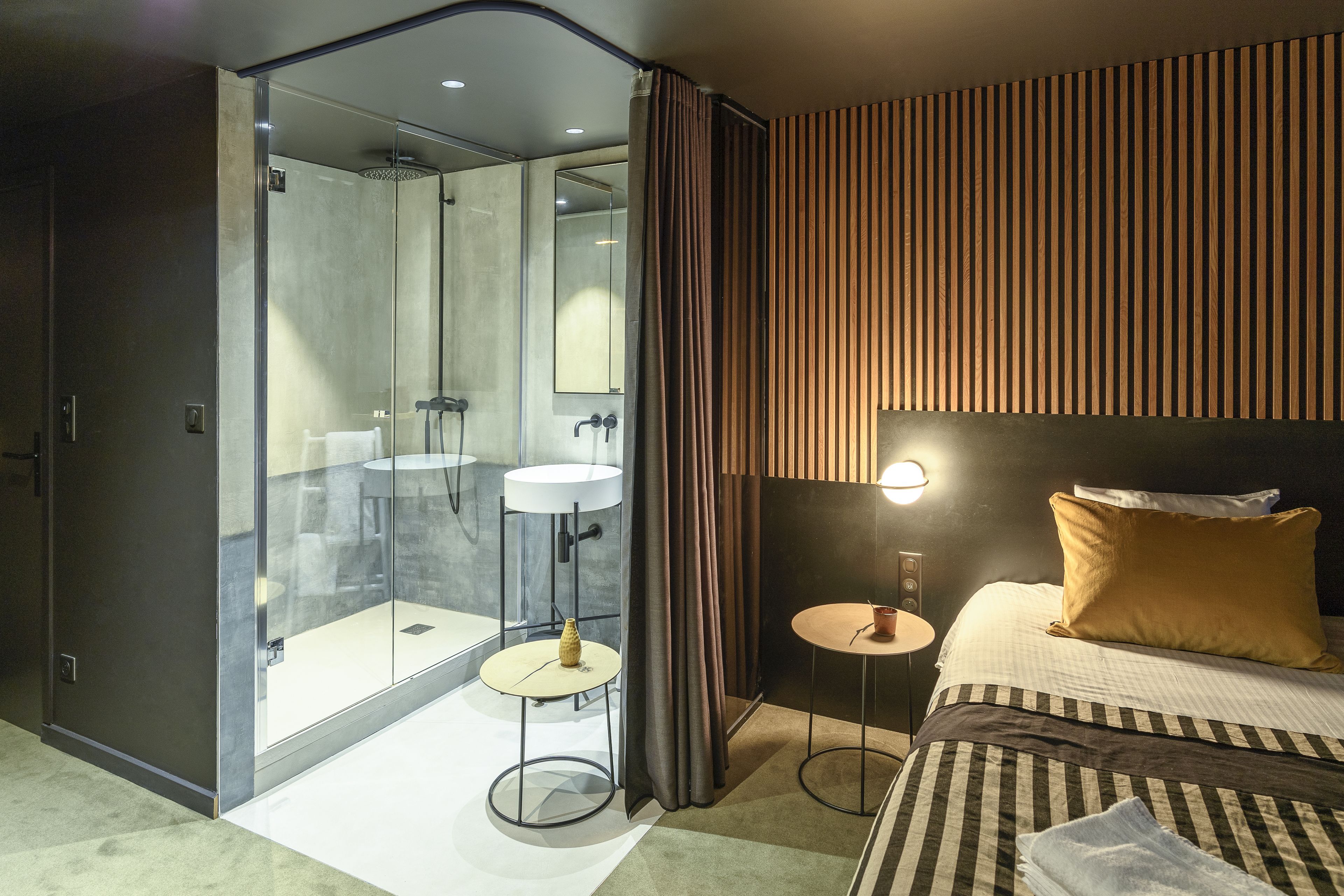 A modern hotel room with a glass-enclosed shower, a bed with striped bedding, and a small bedside table with a lamp.