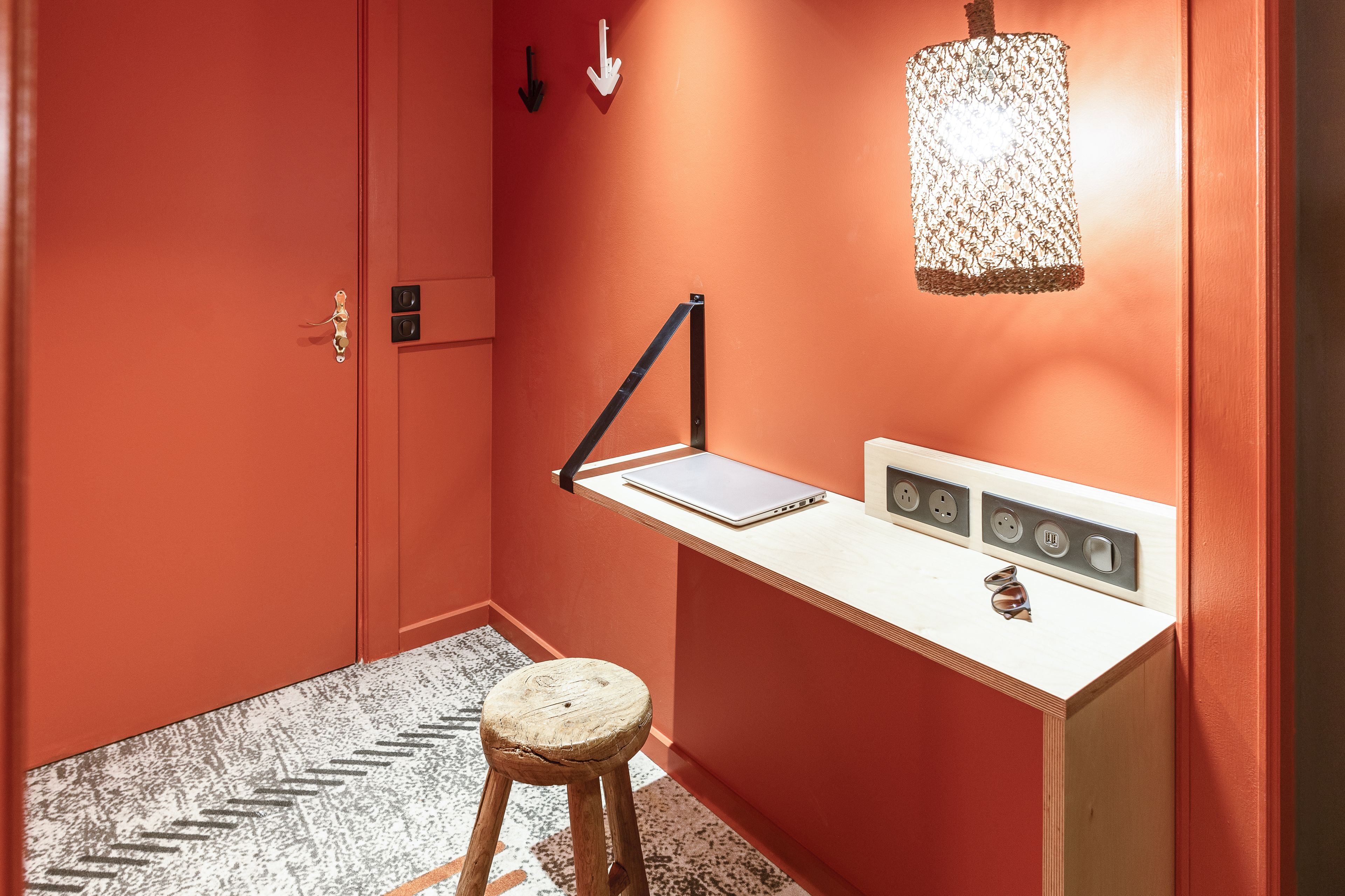 A room with a red wall, a white desk with a laptop, a hanging lamp, and a wooden stool.