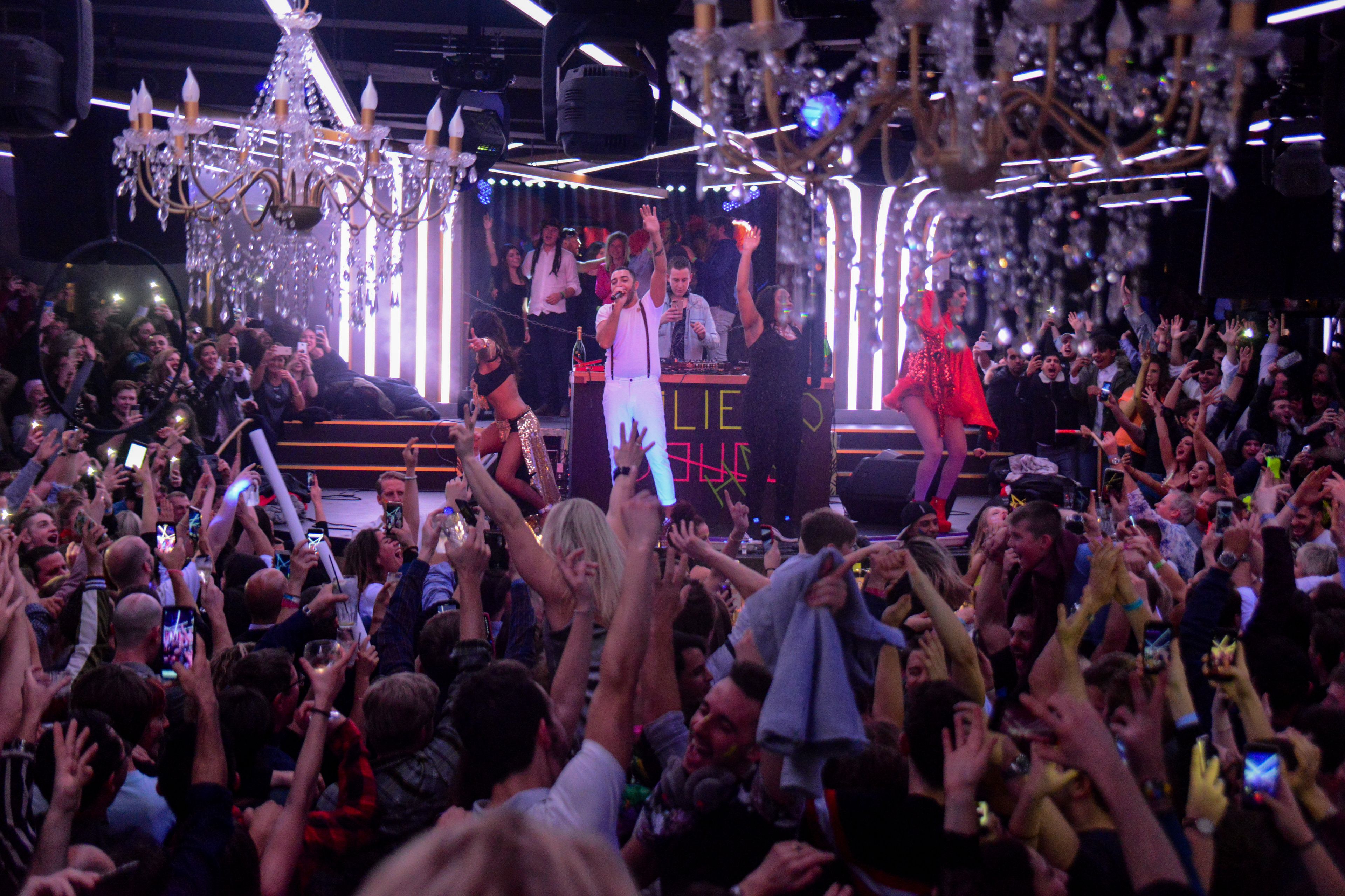 A crowded indoor event with a stage, chandeliers, and a DJ booth.