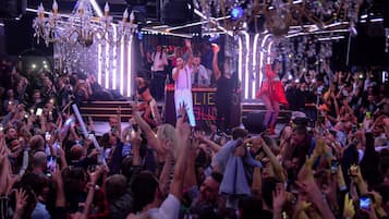 A crowded indoor event with a stage, chandeliers, and a DJ booth.