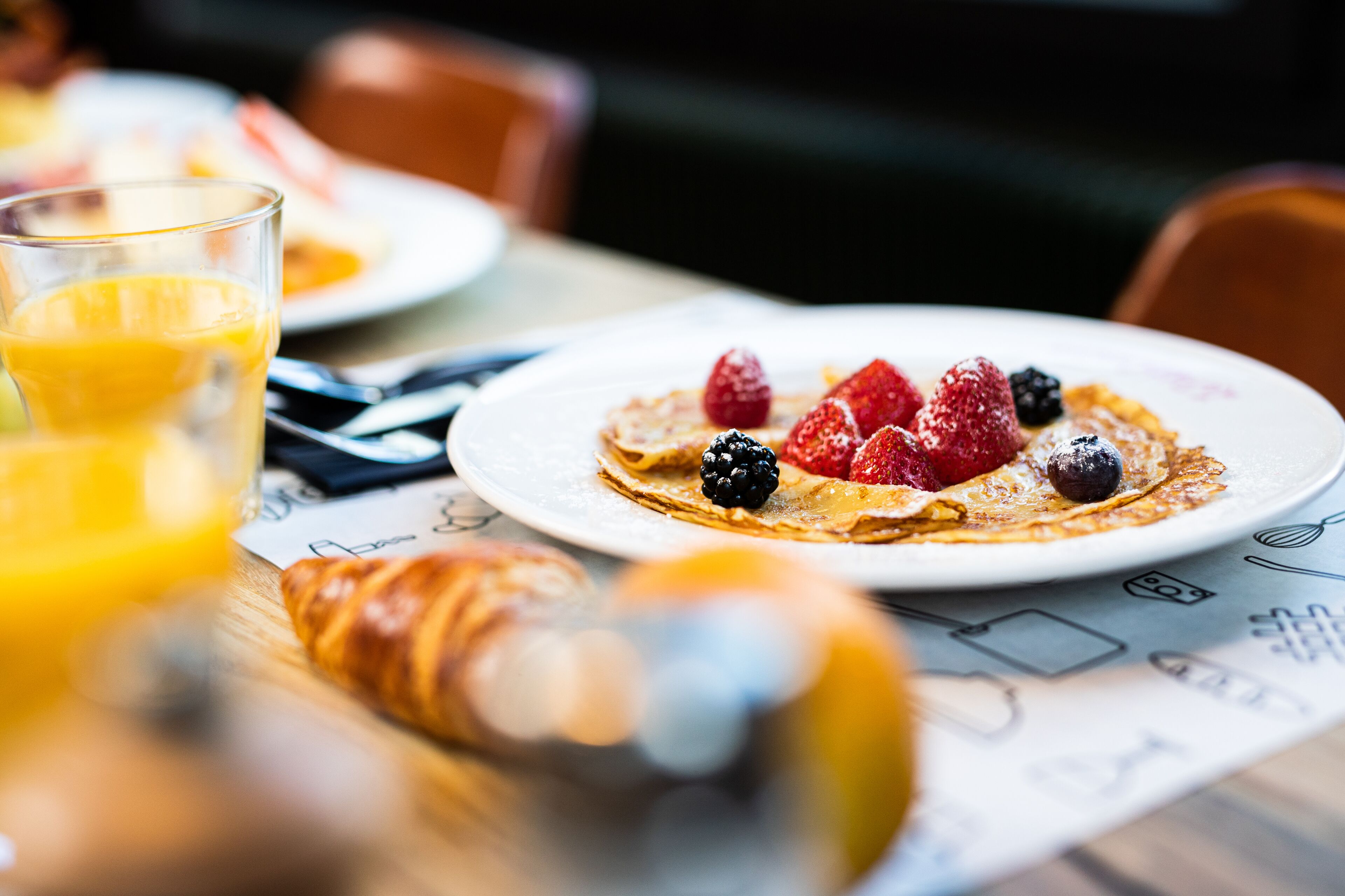 A breakfast setting with a plate of pancakes topped with berries, a glass of orange juice, and a croissant on a table with a menu.