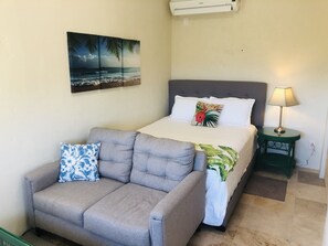 A bedroom with a bed, a grey sofa, a nightstand with a lamp, a wall-mounted air conditioner, and a tropical-themed painting.