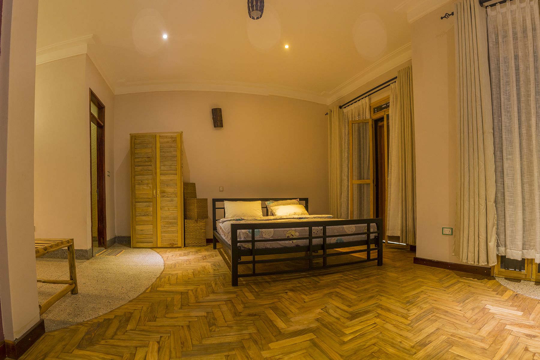 A bedroom with a wooden herringbone floor, a bed with a headboard, a wardrobe, and a window with curtains.