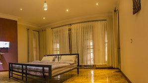 A bedroom with a wooden floor, a bed with a headboard, and a window with curtains. - Dawilderness Holiday Homes (Kampala)