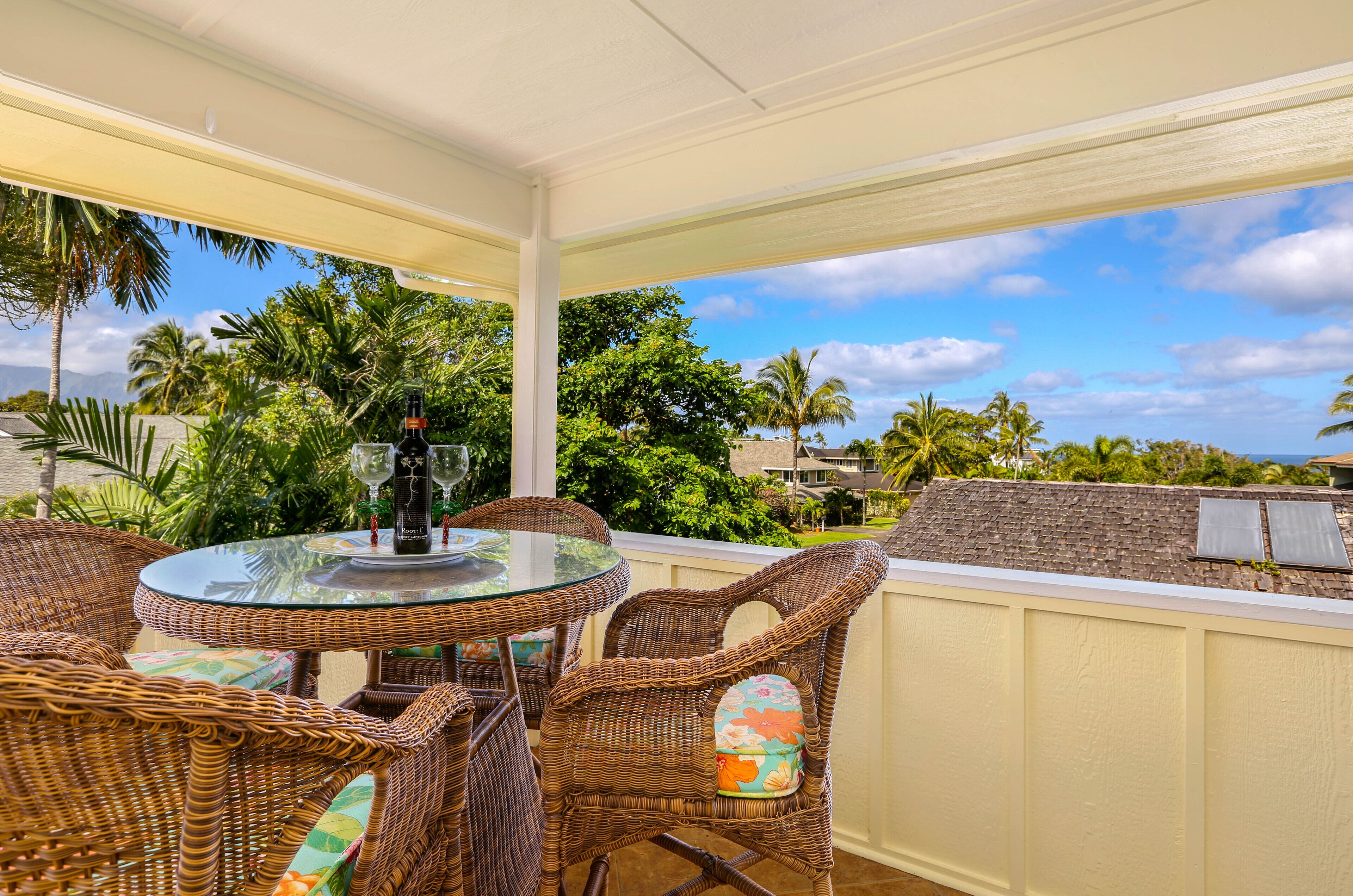 A covered outdoor seating area with wicker furniture and a glass table, overlooking a tropical landscape with palm trees and a clear sky.
