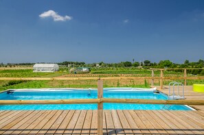 A wooden deck with a swimming pool, a wooden deck, and a view of a green field and a white building in the background. - Tenuta Libò (Alezio)