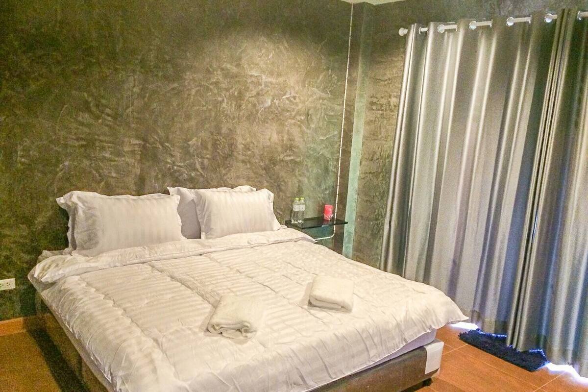 A neatly made bed with white linens, a small bedside table with a bottle and a tissue, and a curtain on a window.