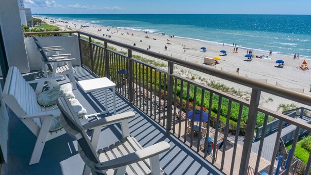A balcony with white chairs and a table, overlooking a beach with umbrellas and people. - Seaside Serenity: Stunning 2-Bedroom Beachfront Condo with Unmatched Ocean Views (Myrtle Beach)