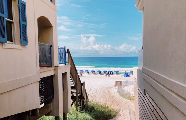Blick auf einen Strand von einem Gebäude mit einem Balkon, einer Holztreppe und einem Zaun, der den Strand begrenzt.
