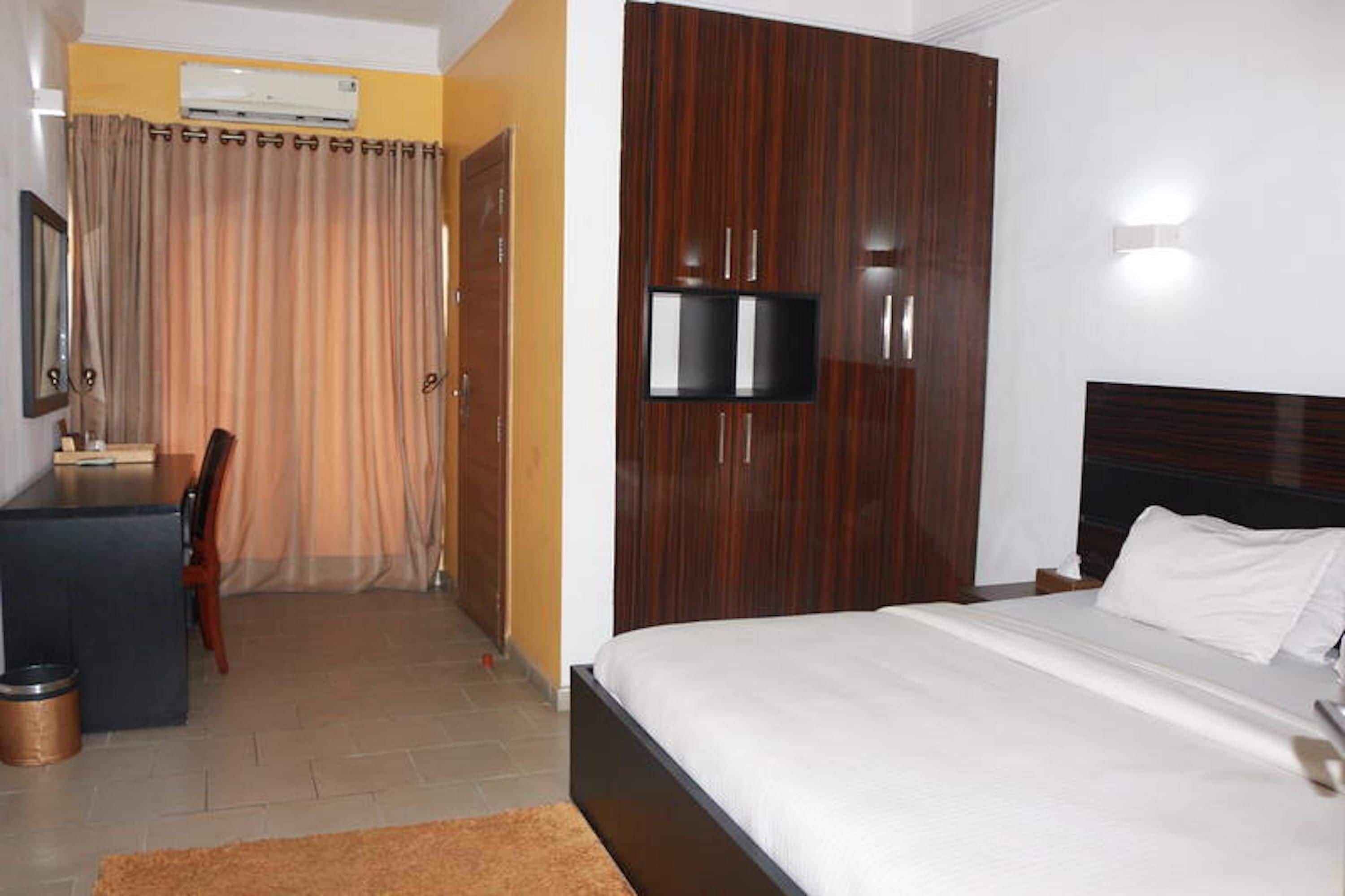 A hotel room with a bed, a TV mounted on a wooden panel, a desk with a chair, and a curtain.