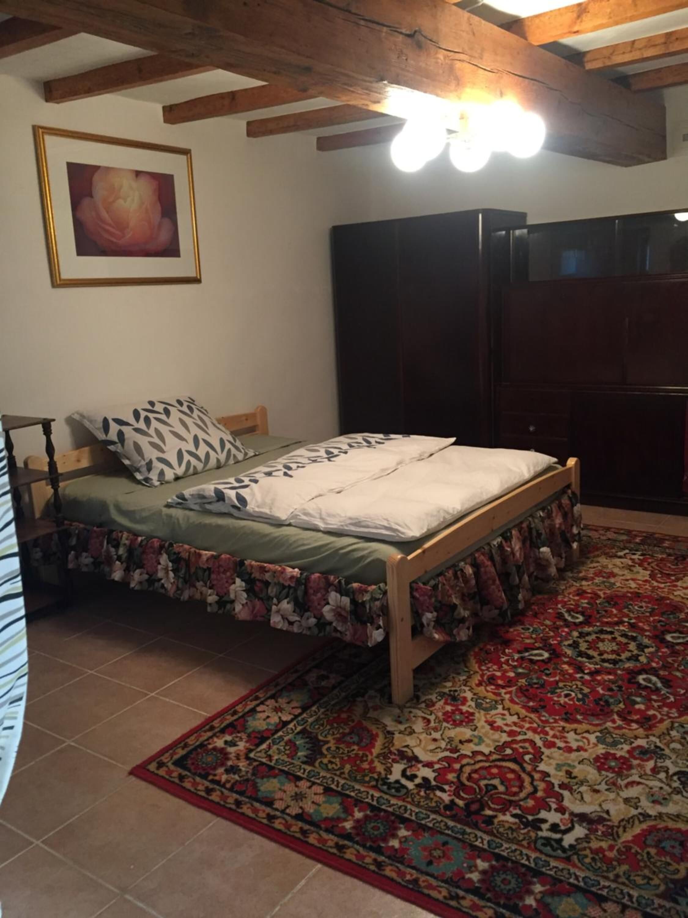 A bedroom with a wooden bed, floral bedspread, a patterned rug, a dark wardrobe, and a framed picture on the wall.