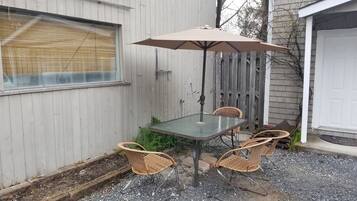 En udendørs terrasse med et glasbord og fire fletstole, en beige parasol og et træhegn i baggrunden.