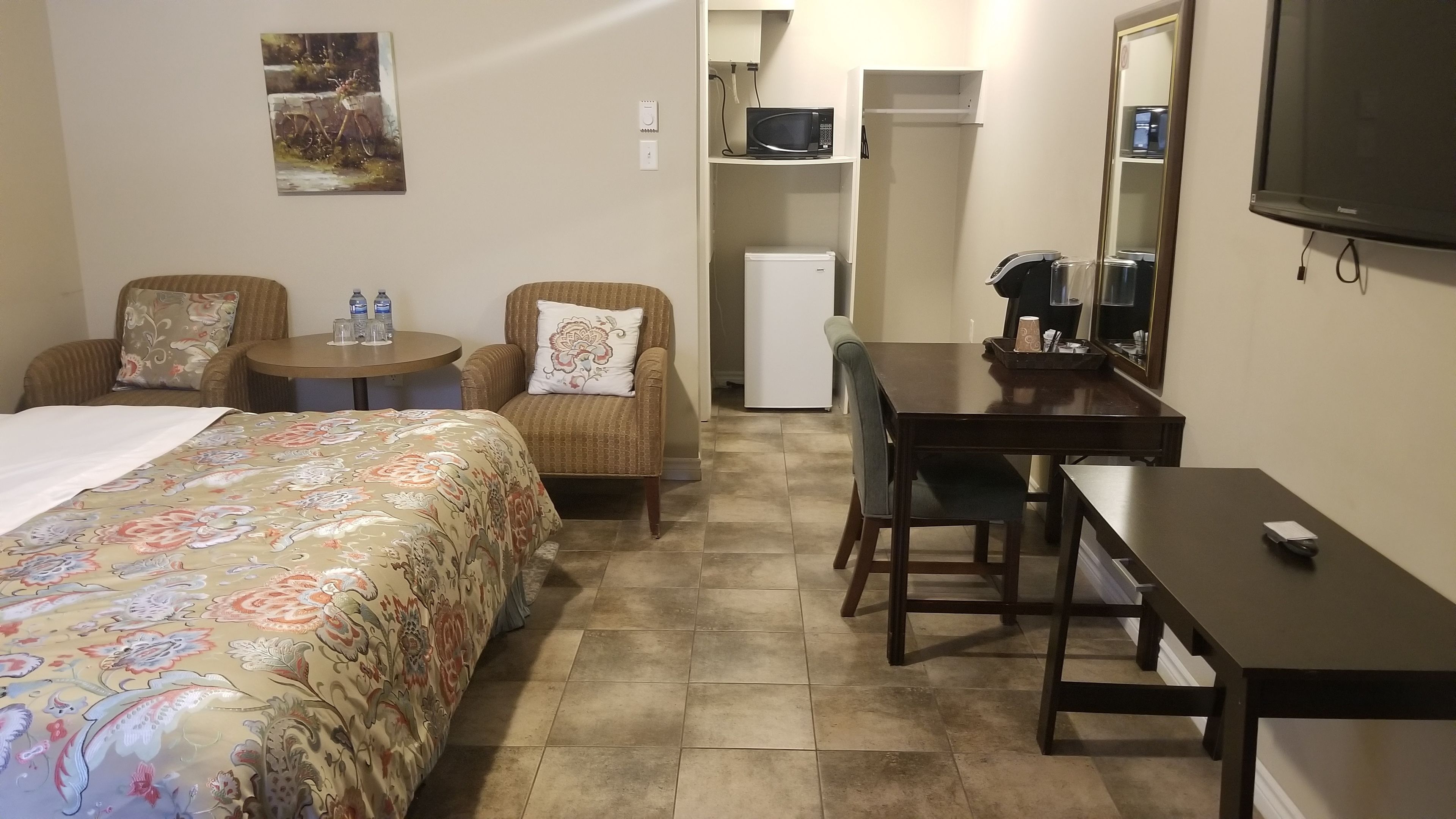 A hotel room with a bed, two armchairs, a small table, a desk, a microwave, a refrigerator, and a television.
