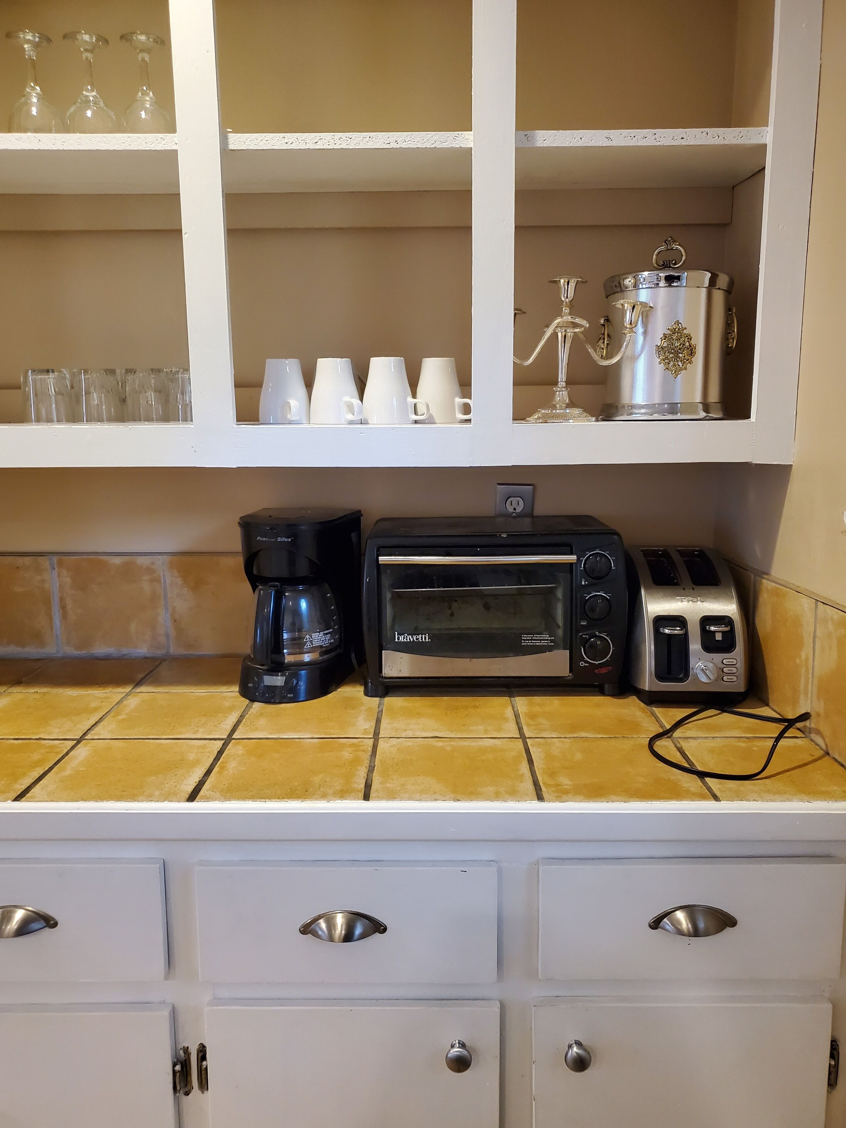 A kitchen with a coffee maker, toaster, and a cabinet with glassware and cups.