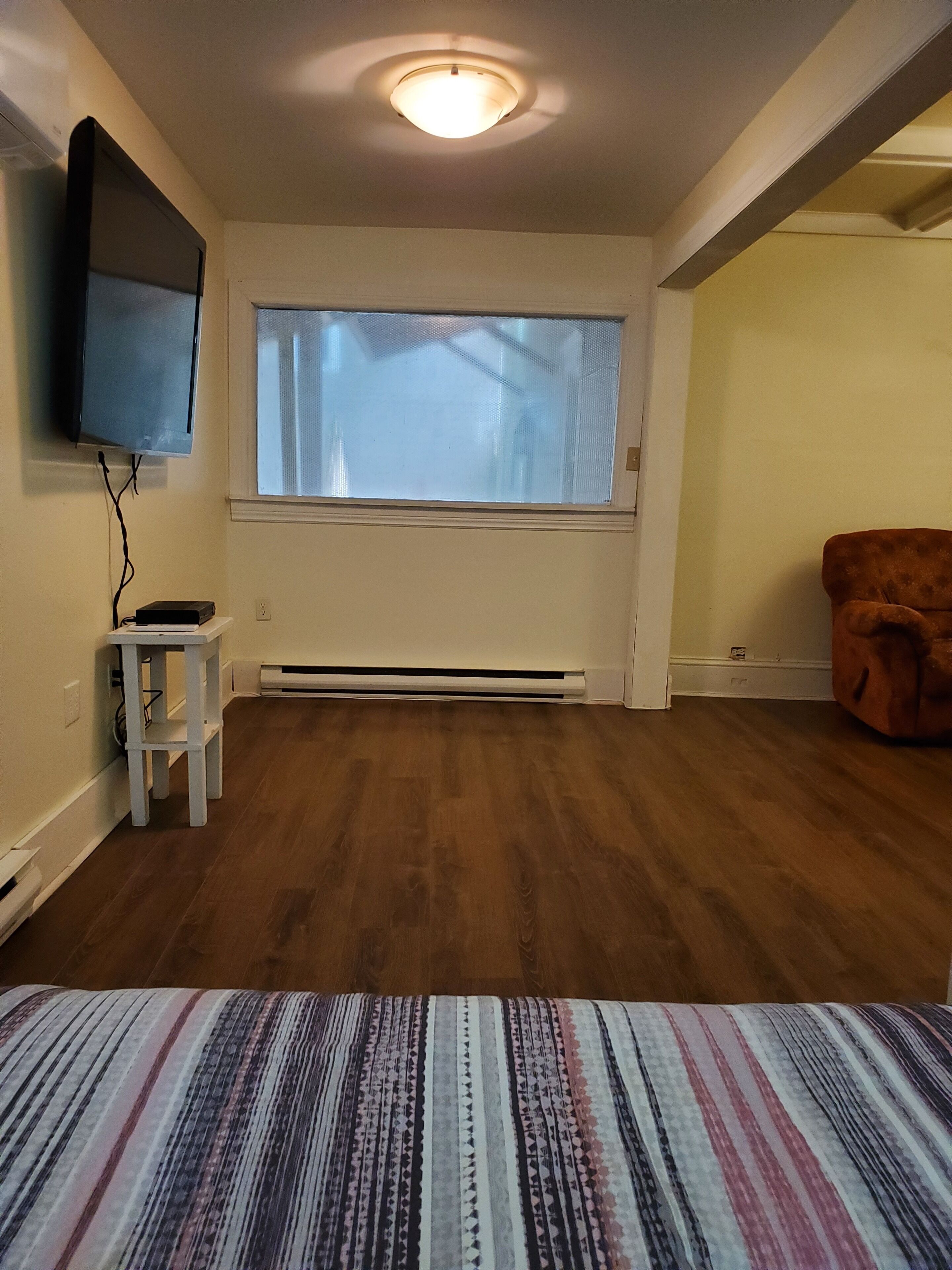 Een kamer met een groot raam, een flatscreen televisie aan de muur, een klein wit tafeltje en een bruine fauteuil.