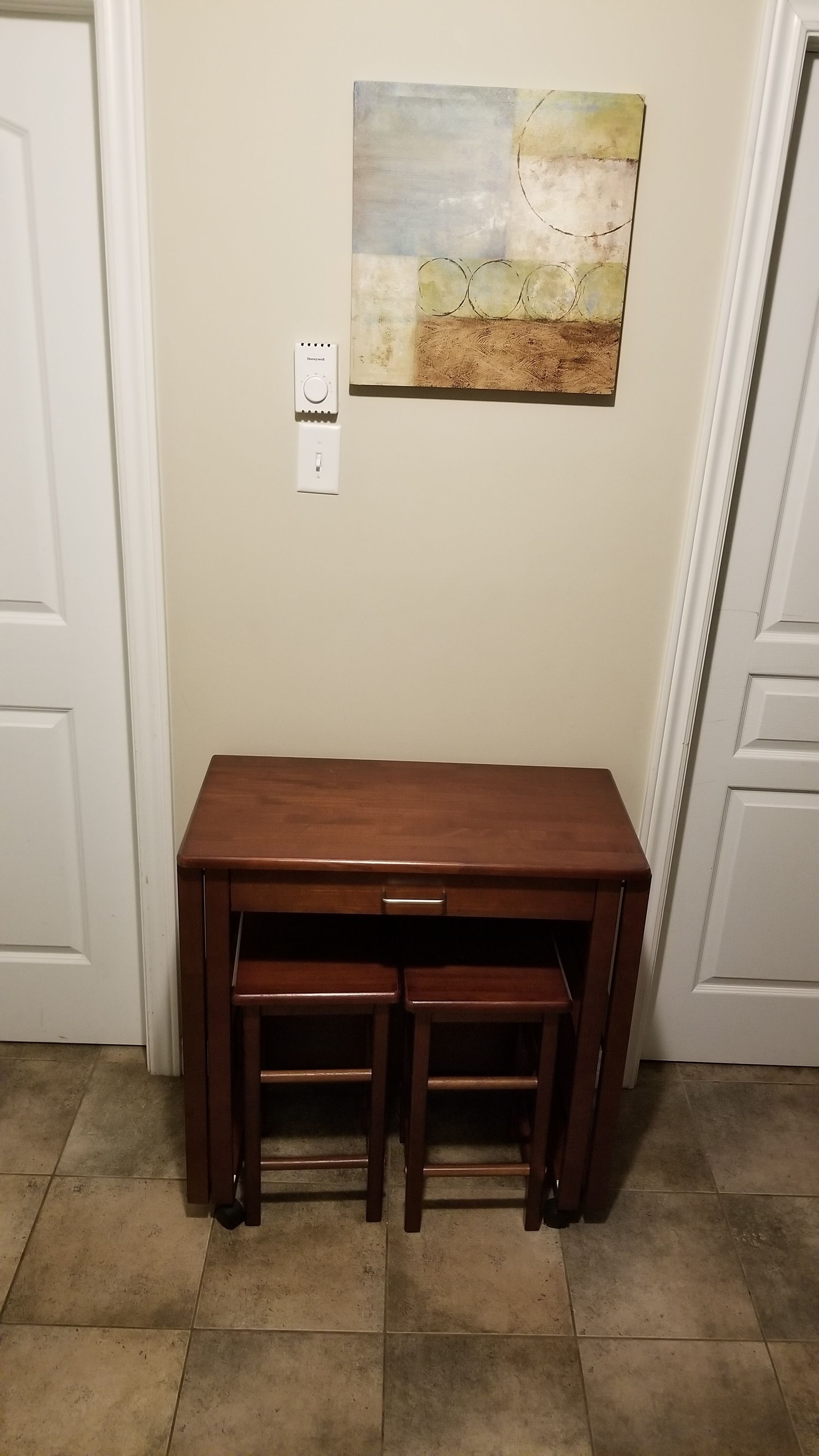 Mesa de madeira com duas cadeiras em um corredor com chão de azulejos e um quadro na parede.