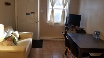 Una habitación con sofá amarillo, televisor negro sobre un mueble bajo, una mesa de comedor con sillas y una ventana con cortinas.