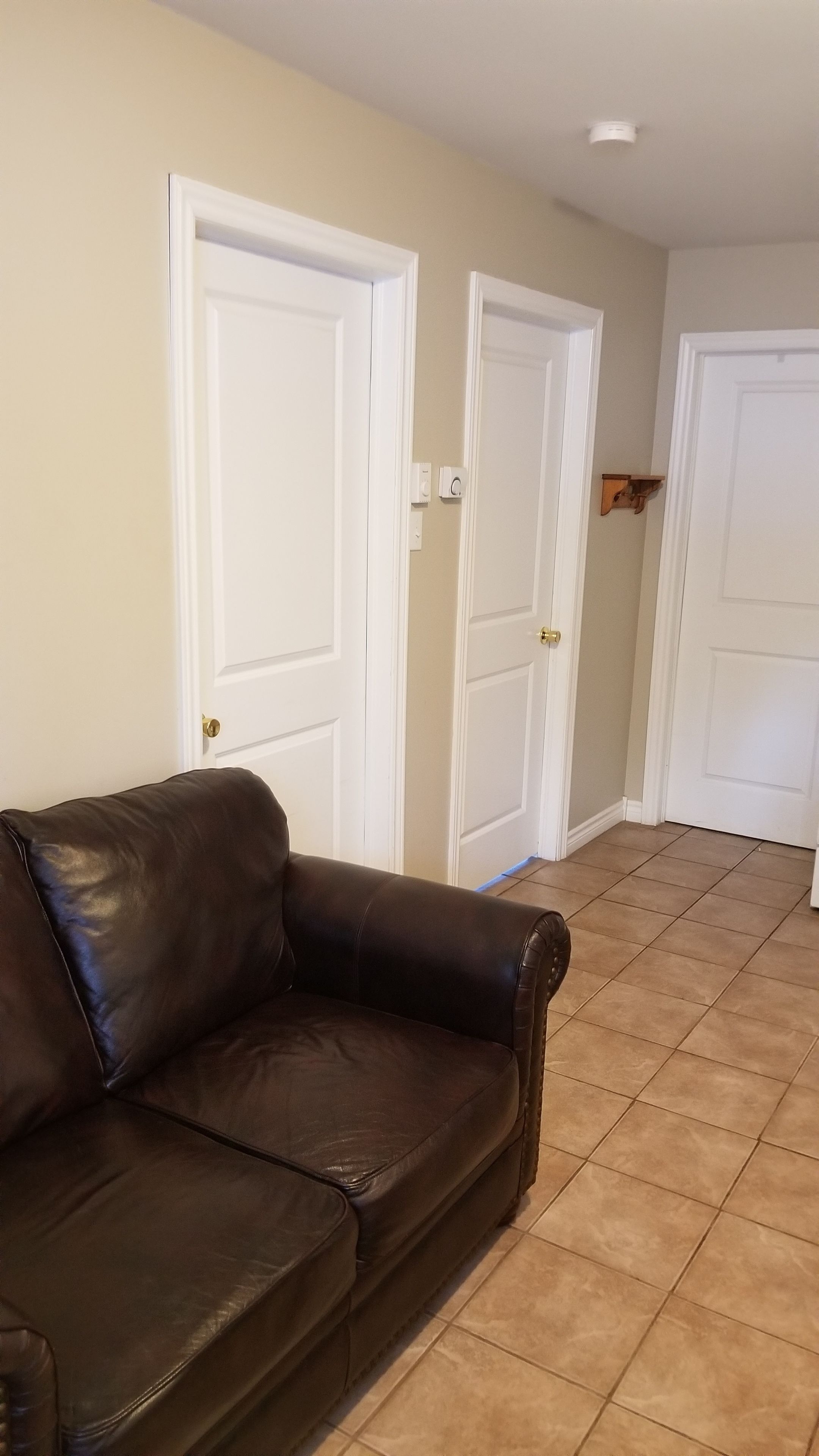 Une pièce avec un canapé en cuir marron, des murs de couleur beige et un sol carrelé. On y trouve trois portes blanches, dont une avec une poignée de porte en laiton.