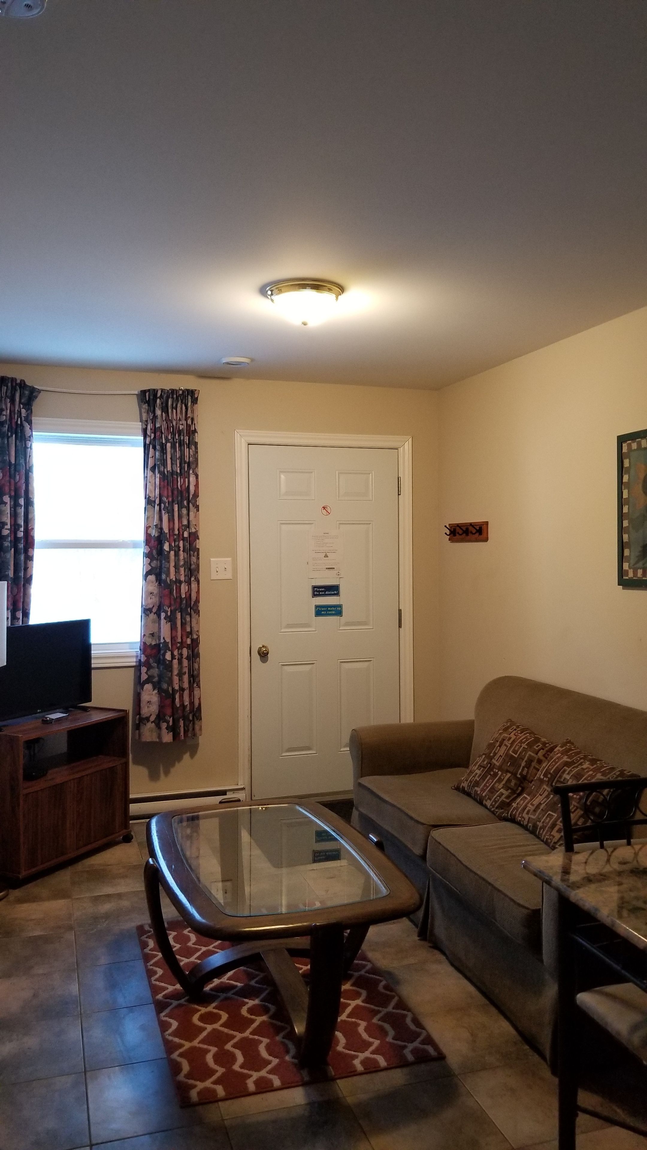 A living room with a glass-top coffee table, a sofa, a TV on a wooden stand, and a door with a sign.
