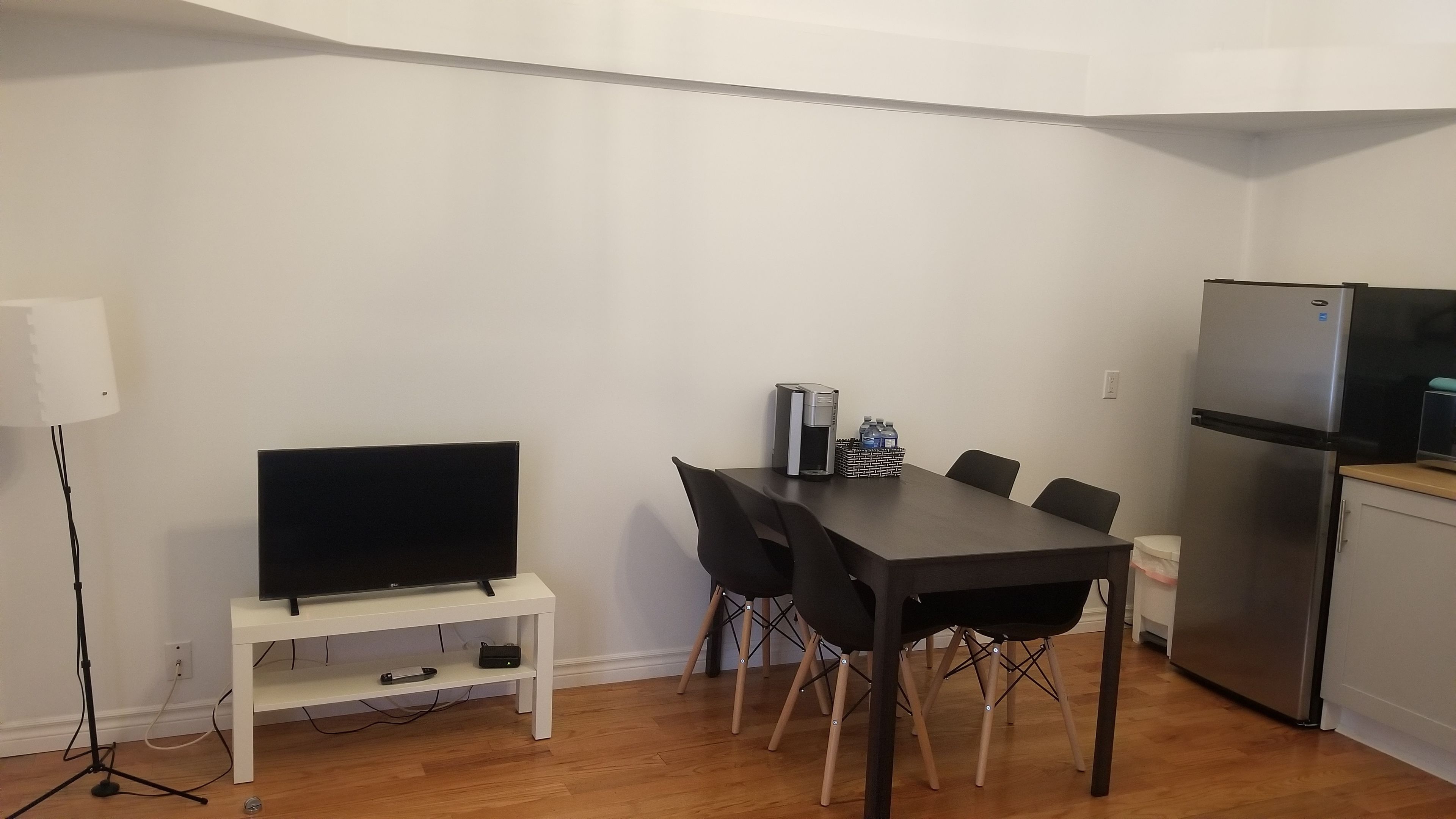 A compact living space with a dining table, chairs, a TV on a stand, a refrigerator, and a coffee maker.