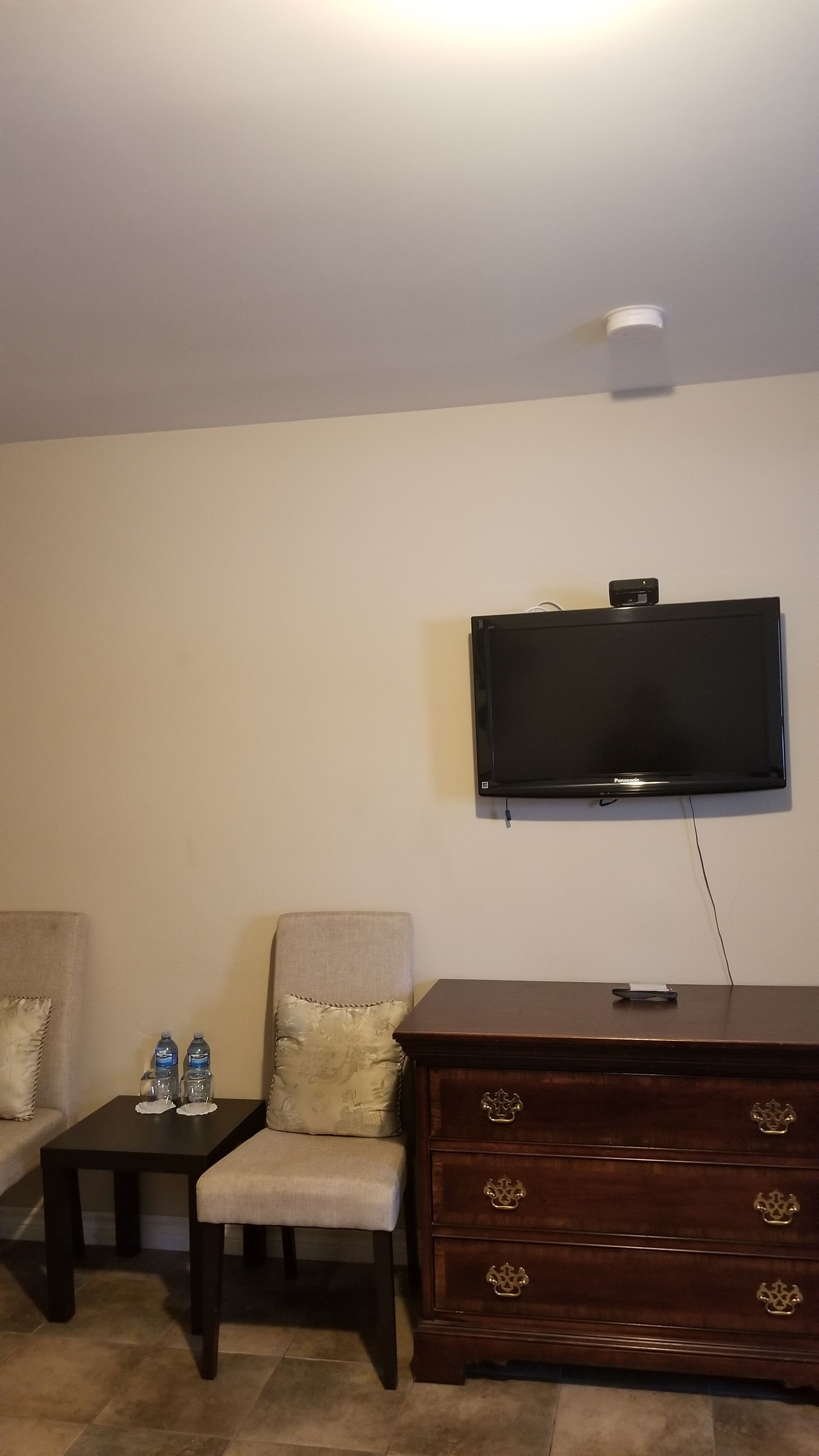 A living room with a flat-screen TV mounted on the wall, a wooden dresser with drawers, a small table with water bottles, and a cushioned armchair.