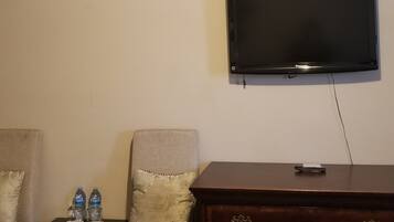 A living room with a flat-screen TV mounted on the wall, a wooden dresser with drawers, a small table with water bottles, and a cushioned armchair.