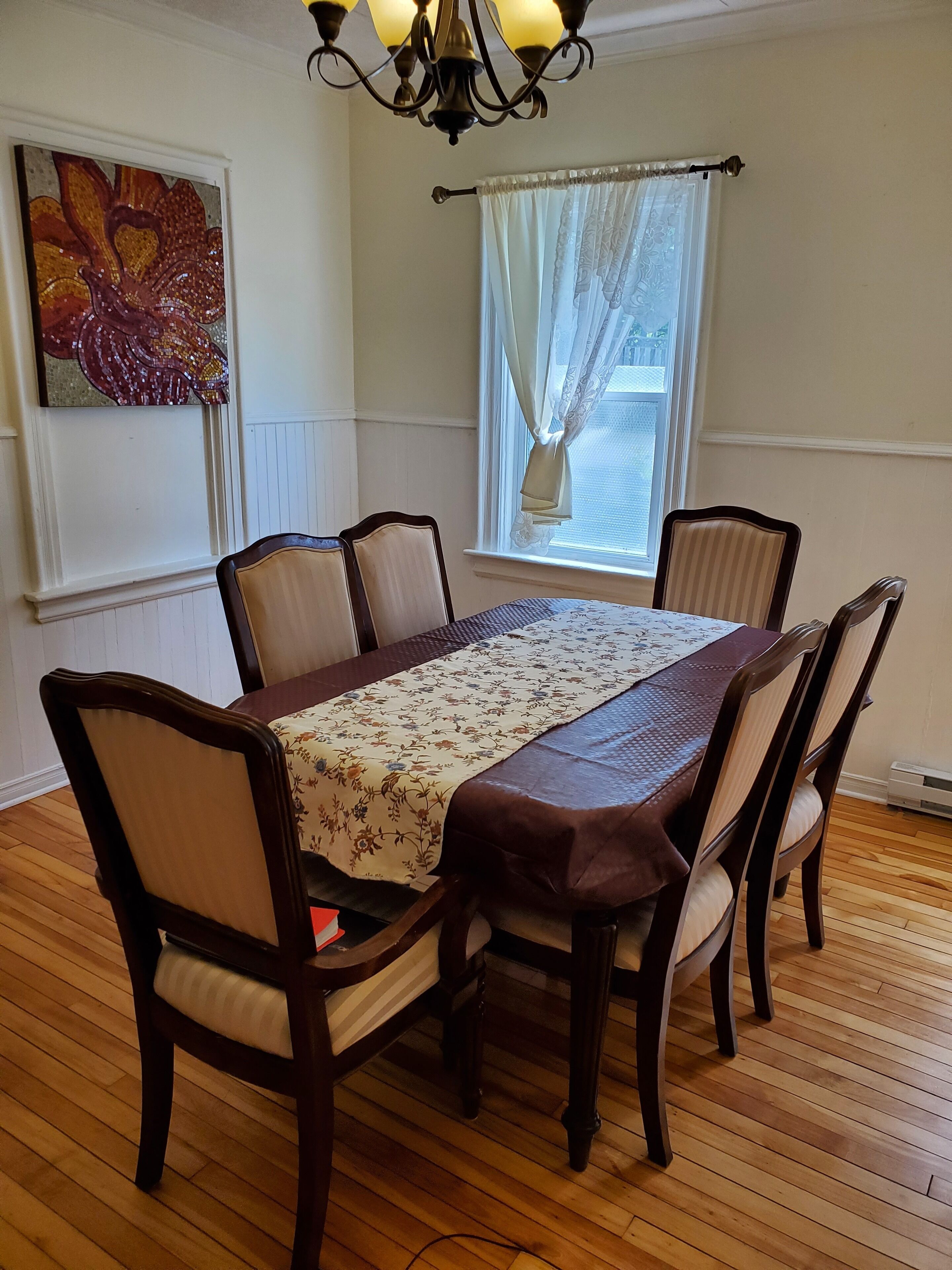 Uma sala de jantar com uma mesa de madeira, seis cadeiras, um candelabro, uma janela com cortinas e um quadro floral na parede.