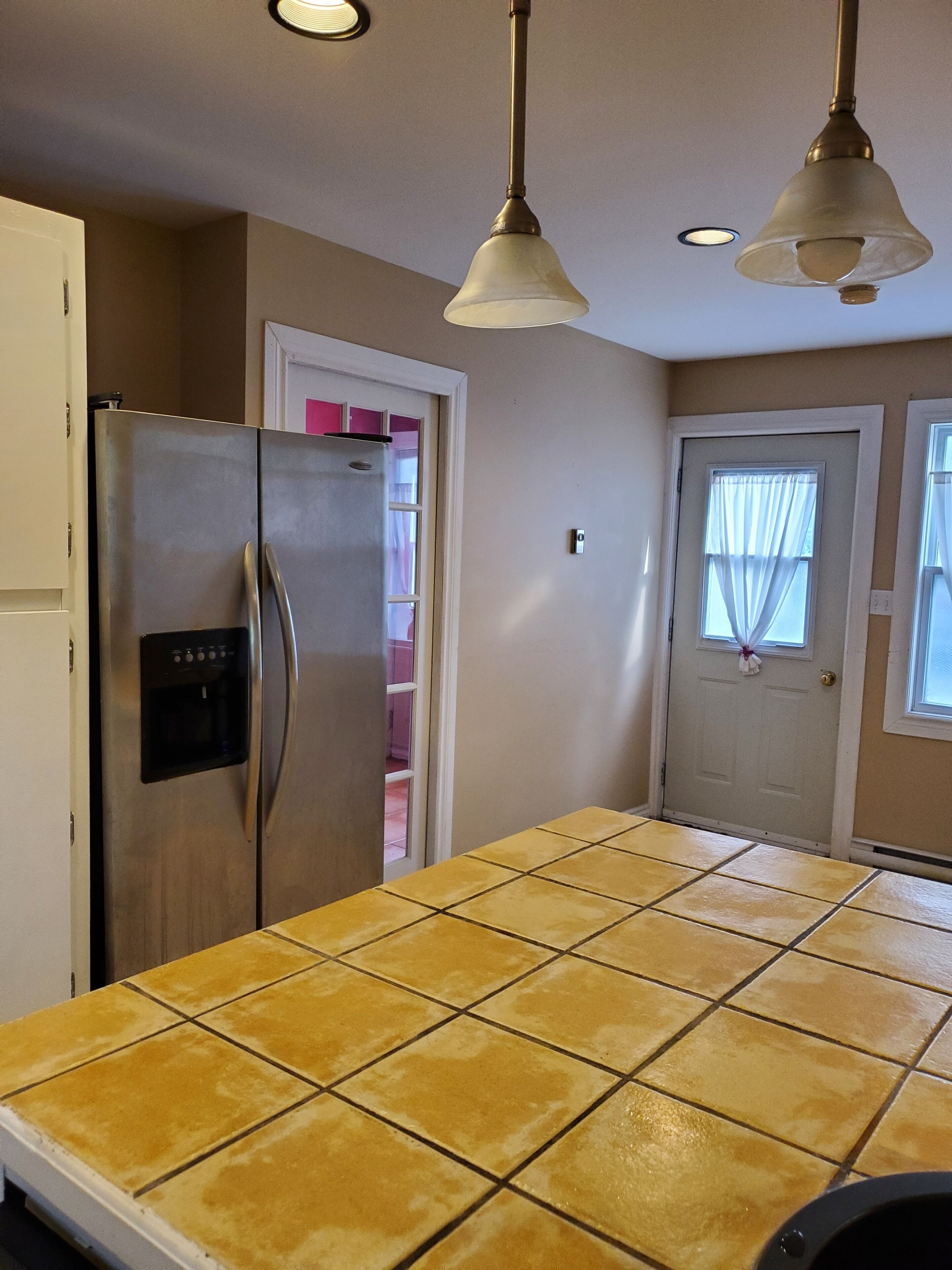 A kitchen with a stainless steel refrigerator, a water dispenser, a tiled countertop, and pendant lights.