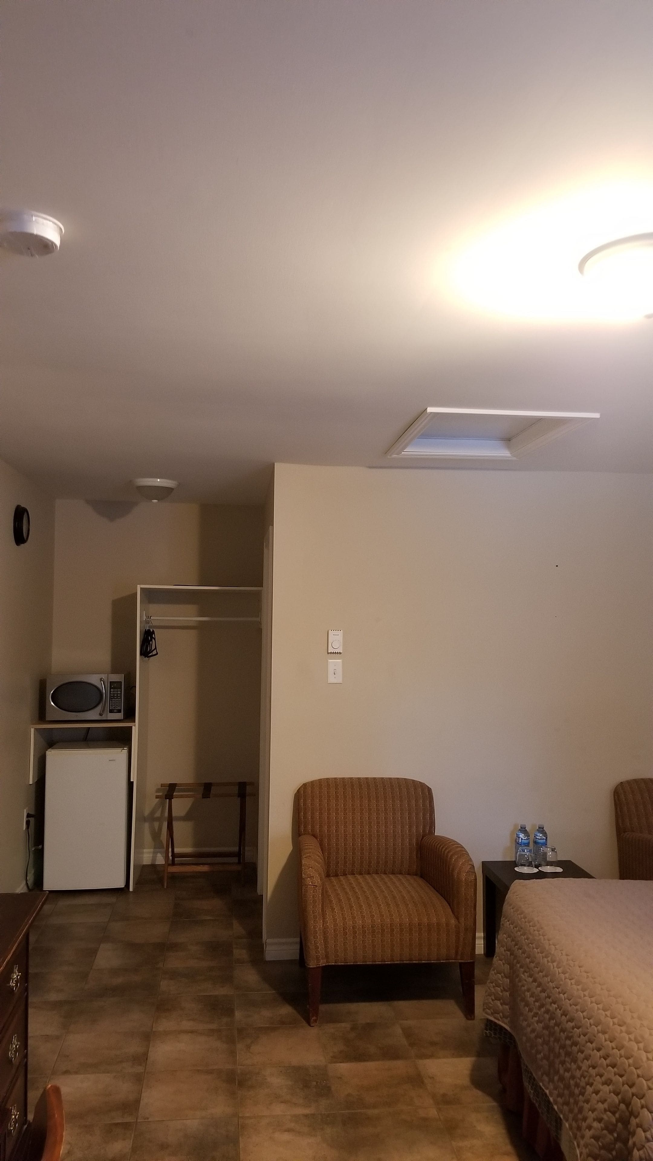 A hotel room with a bed, a chair, a small table with water bottles, a microwave, and a refrigerator.