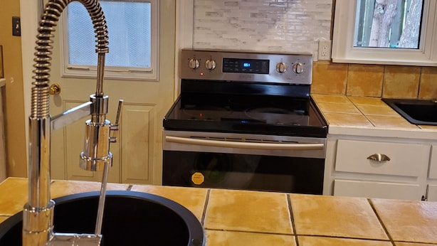 A kitchen with a stainless steel range hood, a built-in oven, a sink with a flexible faucet, and two windows with views of trees. - Rothesay Motel (Rothesay)