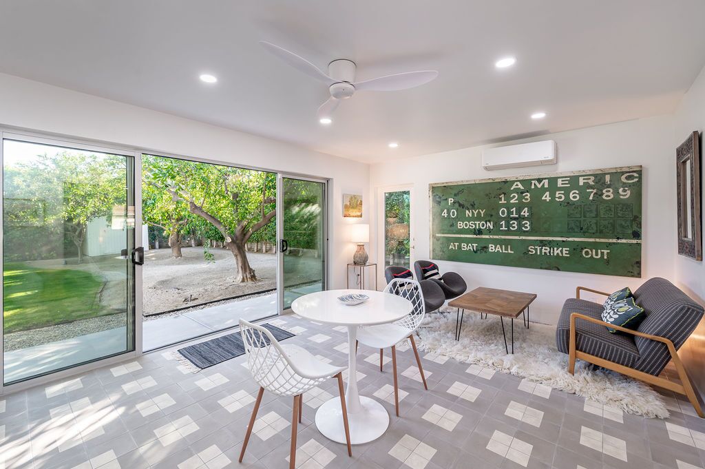 A modern living room with a glass door leading to a garden, a round table with chairs, a sofa, and a wall-mounted air conditioning unit.