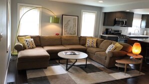 A modern living room with a sectional sofa, a round coffee table, and a small side table with a lamp. There is a kitchen area visible in the background. - Walking distance to downtown Elk Rapids, Elk Lake and dining (Elk Rapids)