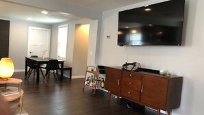 A modern living room with a flat-screen TV mounted on the wall, a wooden cabinet, a dining table with chairs, and a lamp. - Walking distance to downtown Elk Rapids, Elk Lake and dining (Elk Rapids)