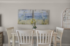 Uma sala de jantar com uma mesa e cadeiras brancas, um quadro de pássaros e flores na parede, e um vaso com planta sobre a mesa.
