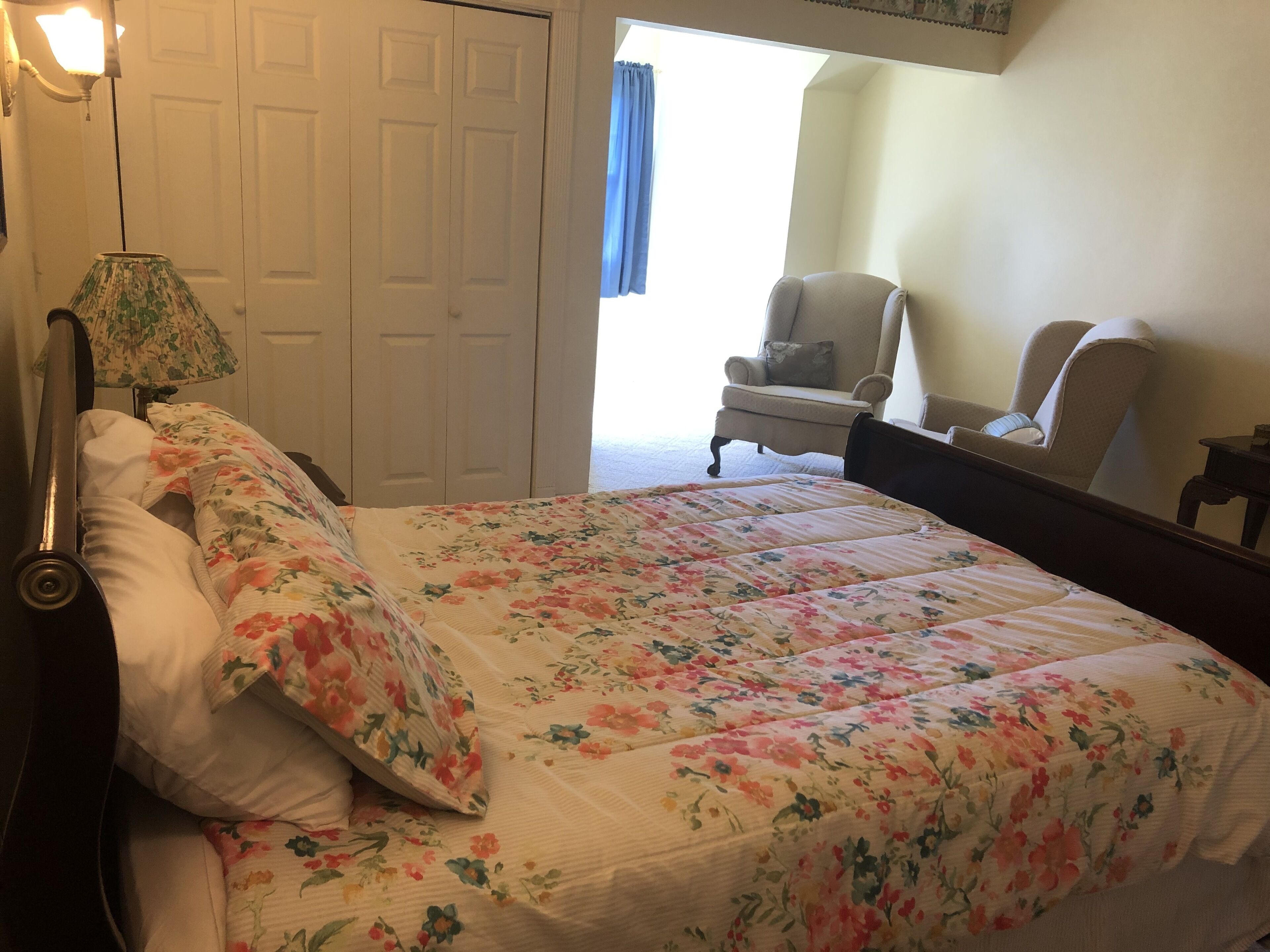A bedroom with a floral bedspread, a bedside lamp, a chair, and a wardrobe.