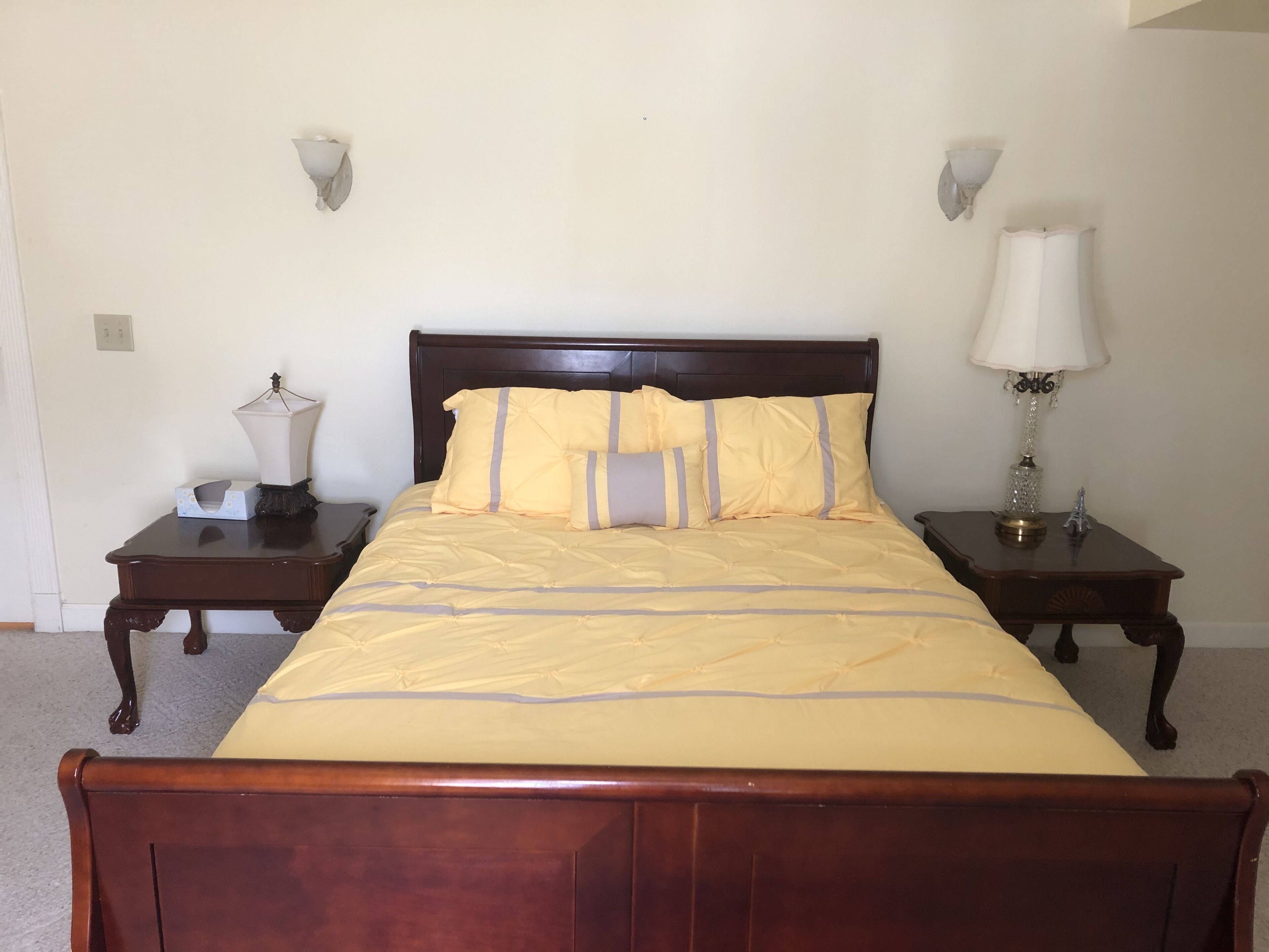 A neatly made bed with a wooden headboard, two bedside tables with lamps, and a plain wall with a light switch.
