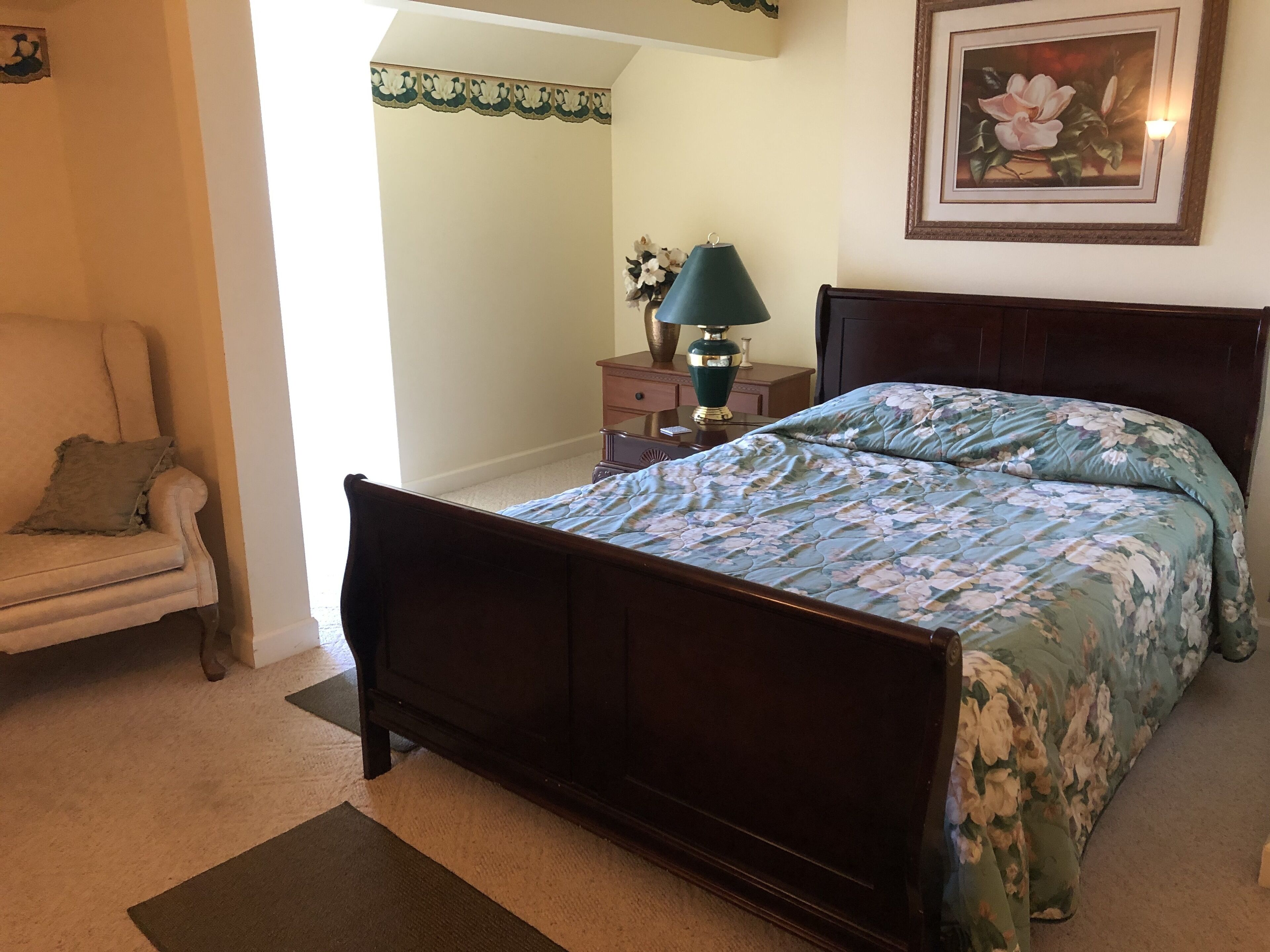 A bedroom with a wooden bed, a chair, a nightstand with a lamp, and a framed picture on the wall.
