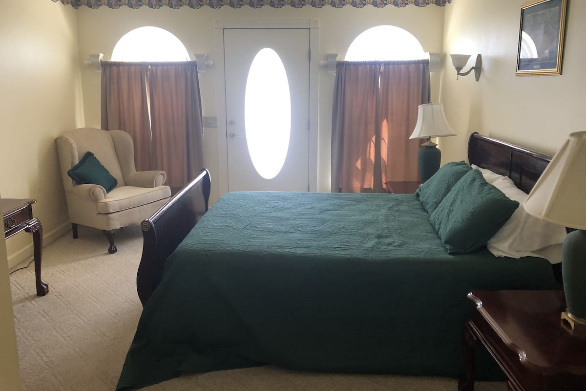 A hotel room with a large bed, a chair, a desk, and two windows with curtains.