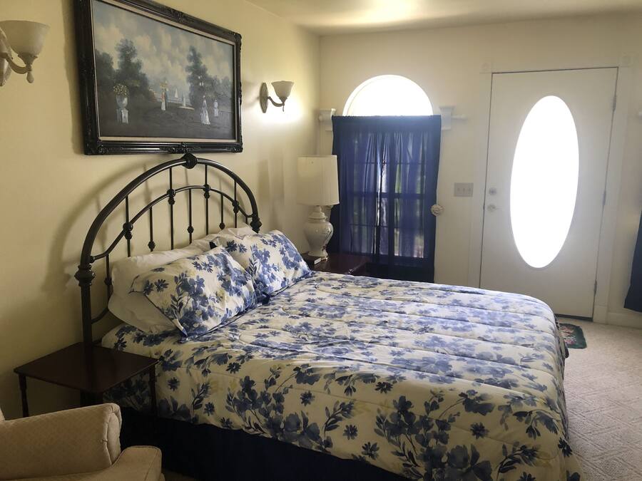 A bedroom with a bed, a nightstand, a lamp, a painting, a window with curtains, and a door.