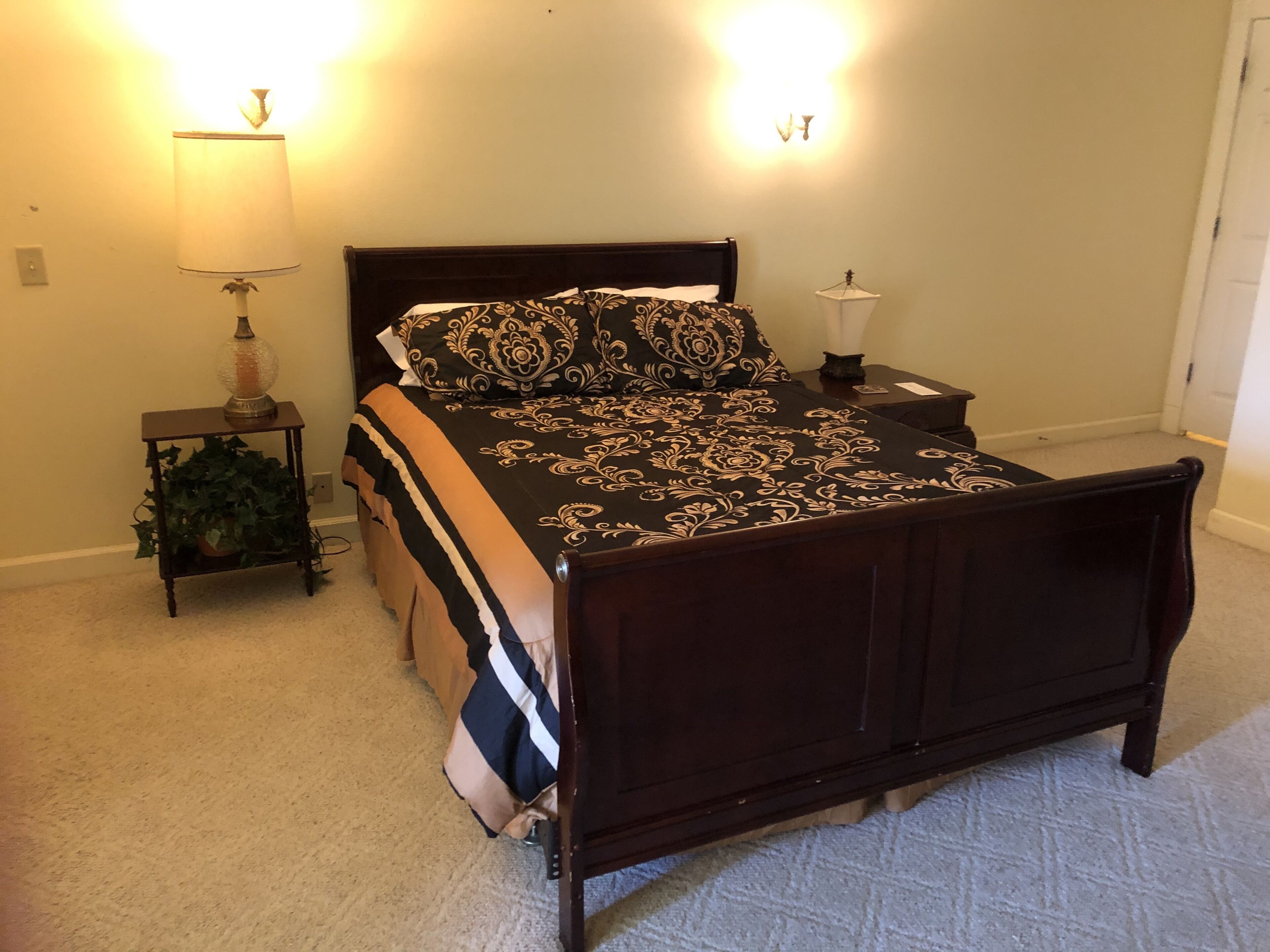 A bedroom with a dark wooden bed, patterned bedding, two bedside lamps, and a small table with a plant.