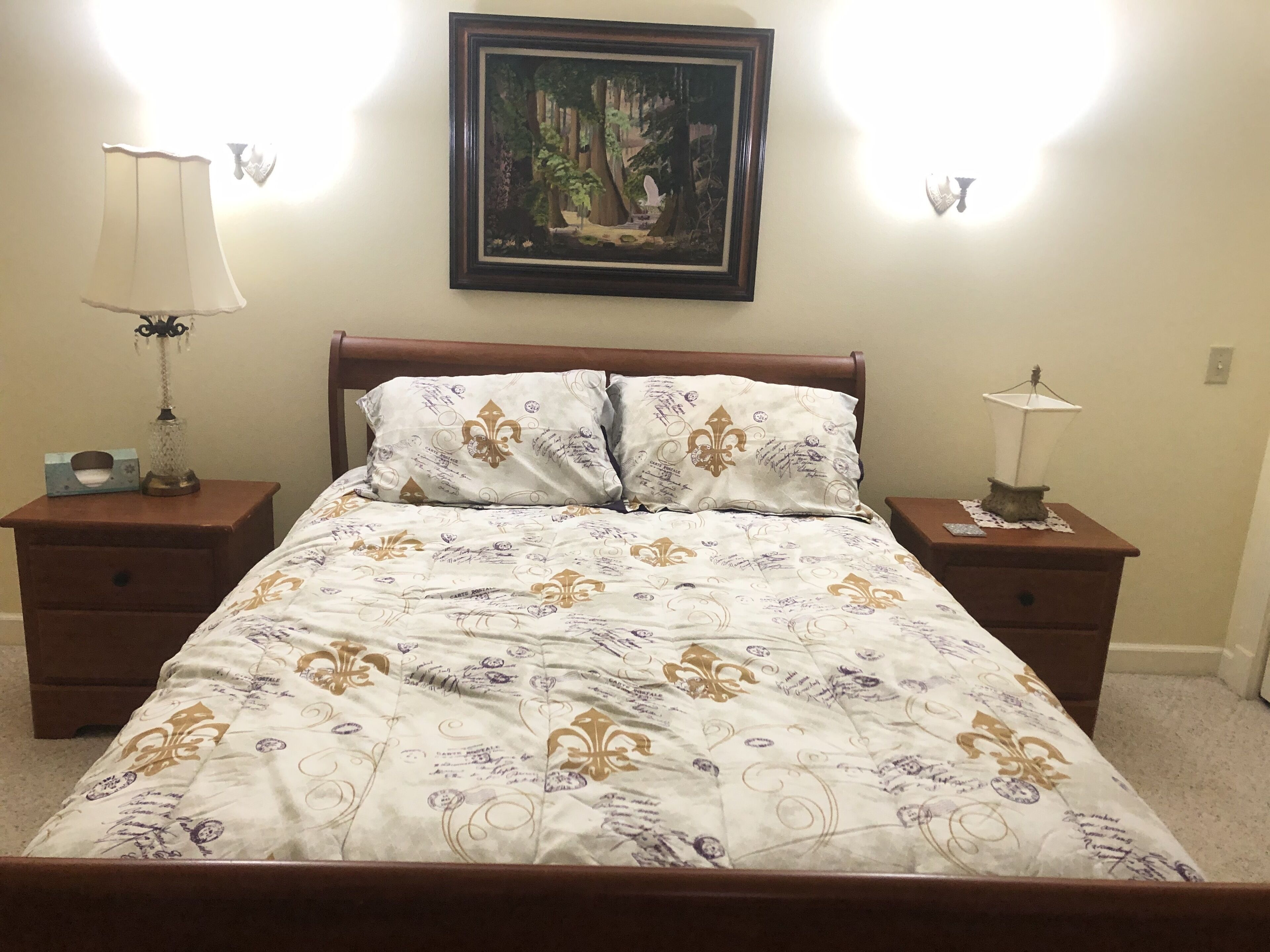 A bedroom with a bed, two nightstands, a framed painting, and two wall-mounted lamps.