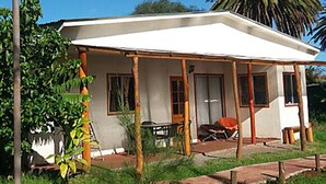 A small, single-story house with a white exterior, a porch with wooden pillars, and a large palm tree in the background.